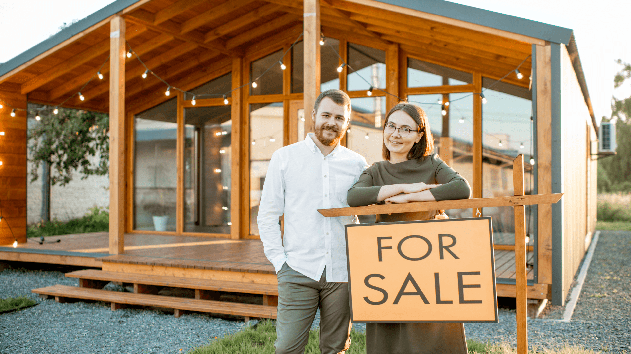 Photo of a couple standing outside of a house holding a "For Sale" sign.